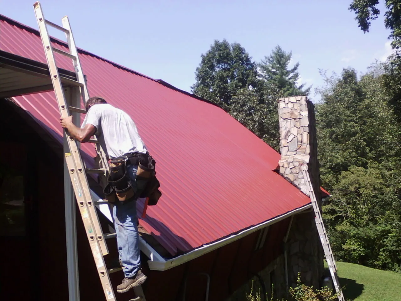 Roofer on a ladder beside a red metal roof and stone chimney — Soffit & Fascia Repair by Roofing Brothers in Pala